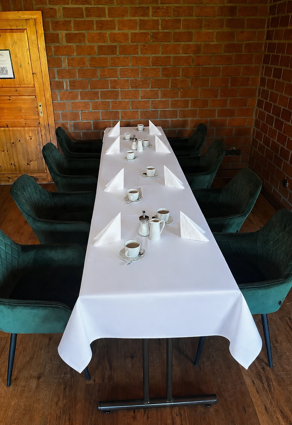Lange eingedeckte Tafel mit Geschirr und Tassen vor Backsteinwand, gepolsterte grüne Stühle